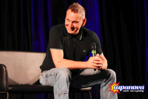 Christopher Eccleston smiling on stage at Supanova 2022 - Melbourne. He is holding a microphone and looking off to the audience.