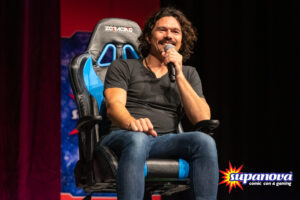 Luke Arnold on stage at Supanova 2021 - Gold Coast, holding a microphone and smiling.